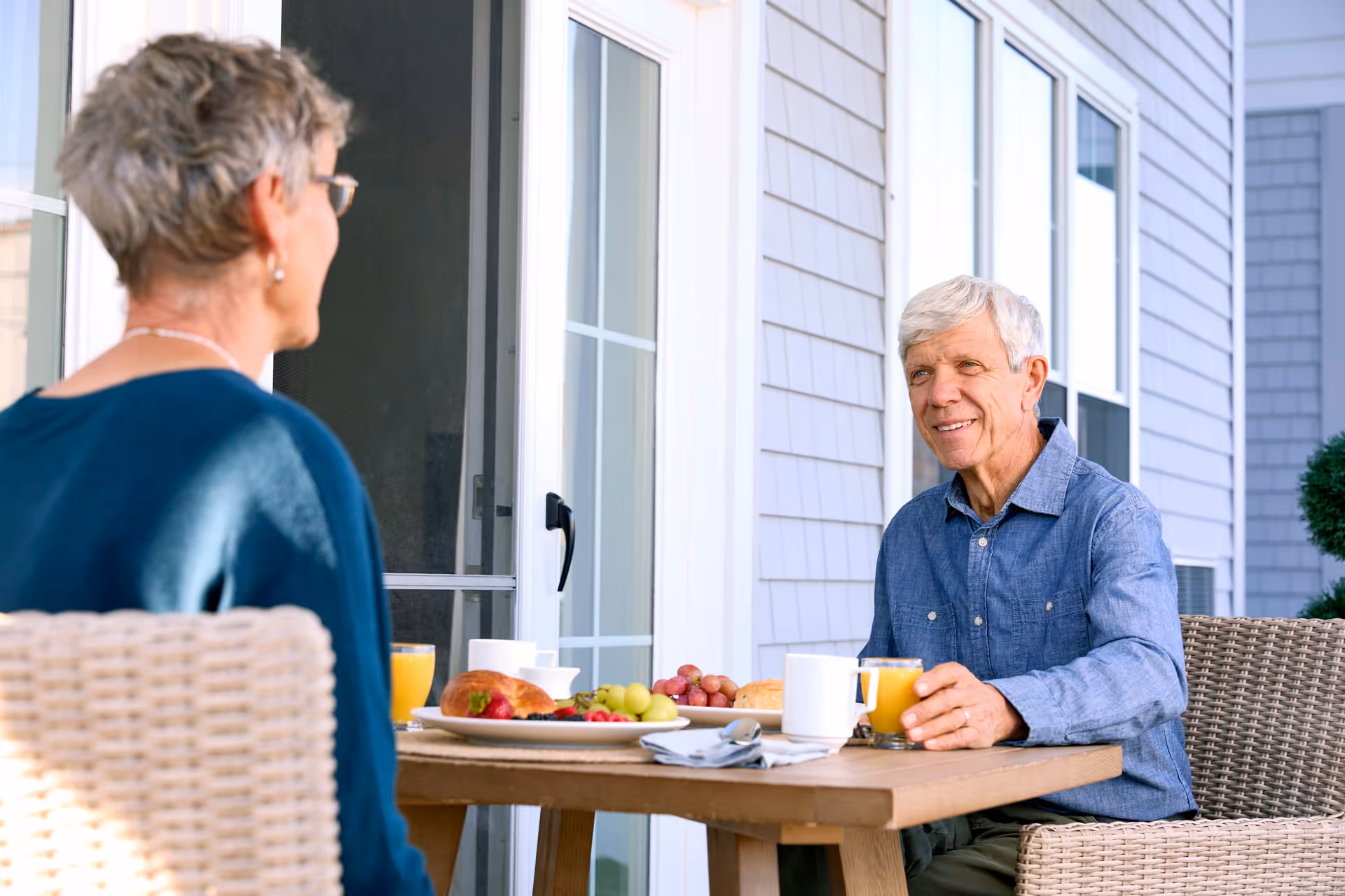 An elderly man and woman sitting at a wooden table outdoors on a patio, enjoying breakfast with plates of fruit, croissants, coffee cups, and glasses of orange juice. The man is smiling and holding a glass of orange juice, while the woman is seen from behind. The background shows the exterior siding and windows of a building.