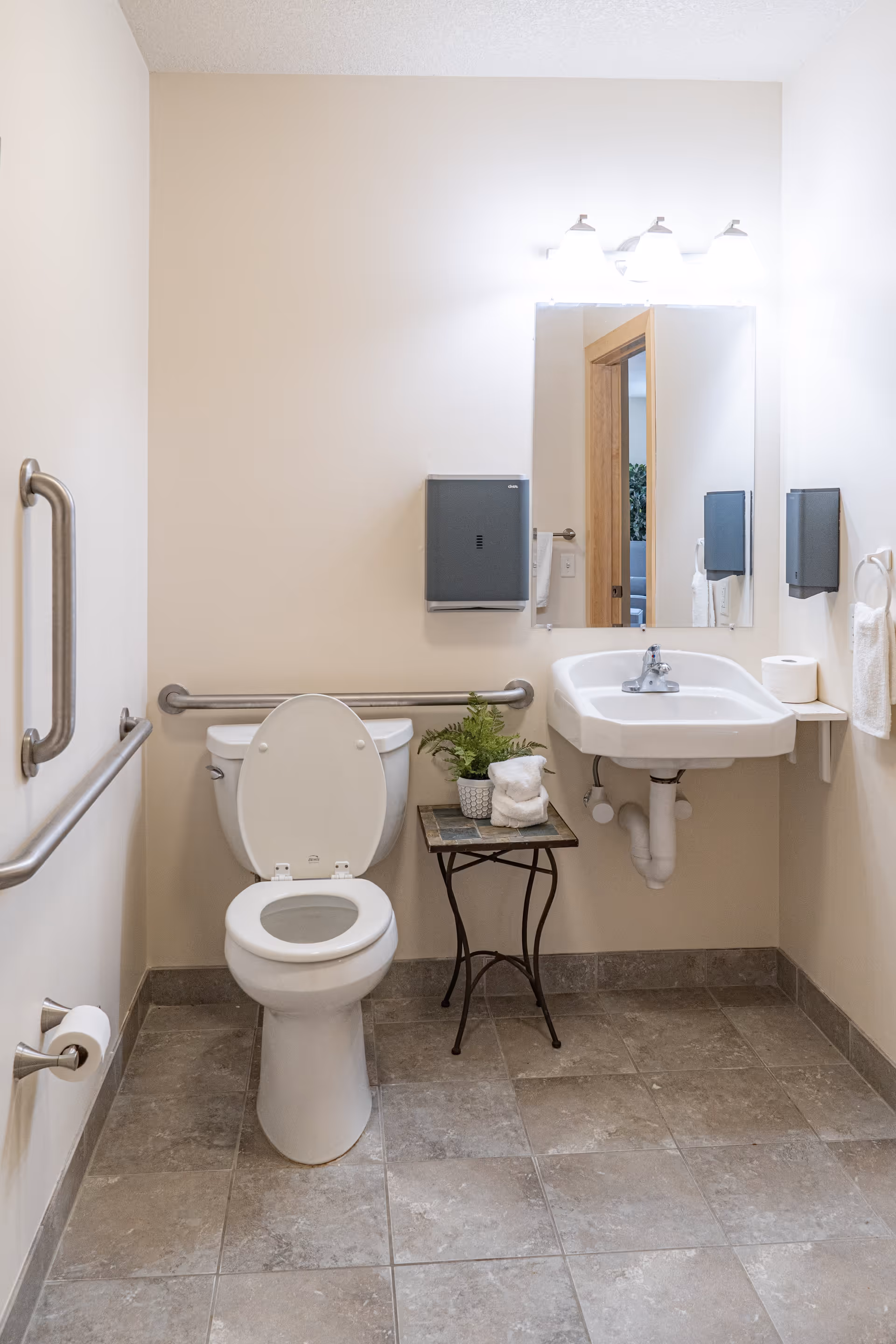 A clean and accessible bathroom with a white toilet featuring grab bars on both sides, a white sink with a mirror above it, a small table holding a potted plant and rolled towels, and a paper towel dispenser mounted on the wall.