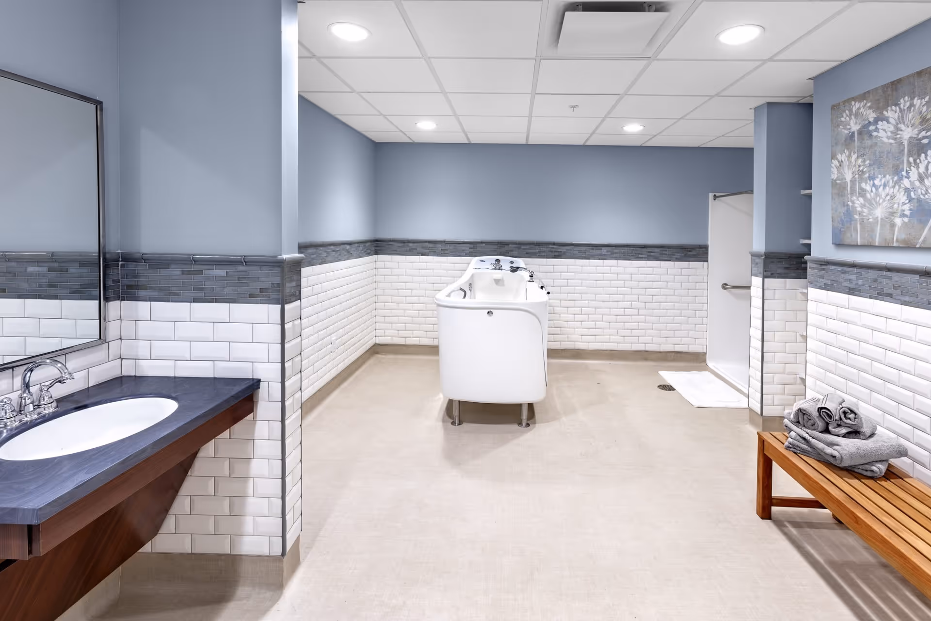 A clean and spacious assisted living bathroom with light blue walls and white subway tiles halfway up the walls. The room features a white walk-in bathtub in the center, a sink with a dark countertop and a large mirror on the left, a wooden bench with neatly folded gray towels on the right, and a walk-in shower with a grab bar in the back right corner. The ceiling has recessed lighting, and there is a decorative floral painting on the right wall.