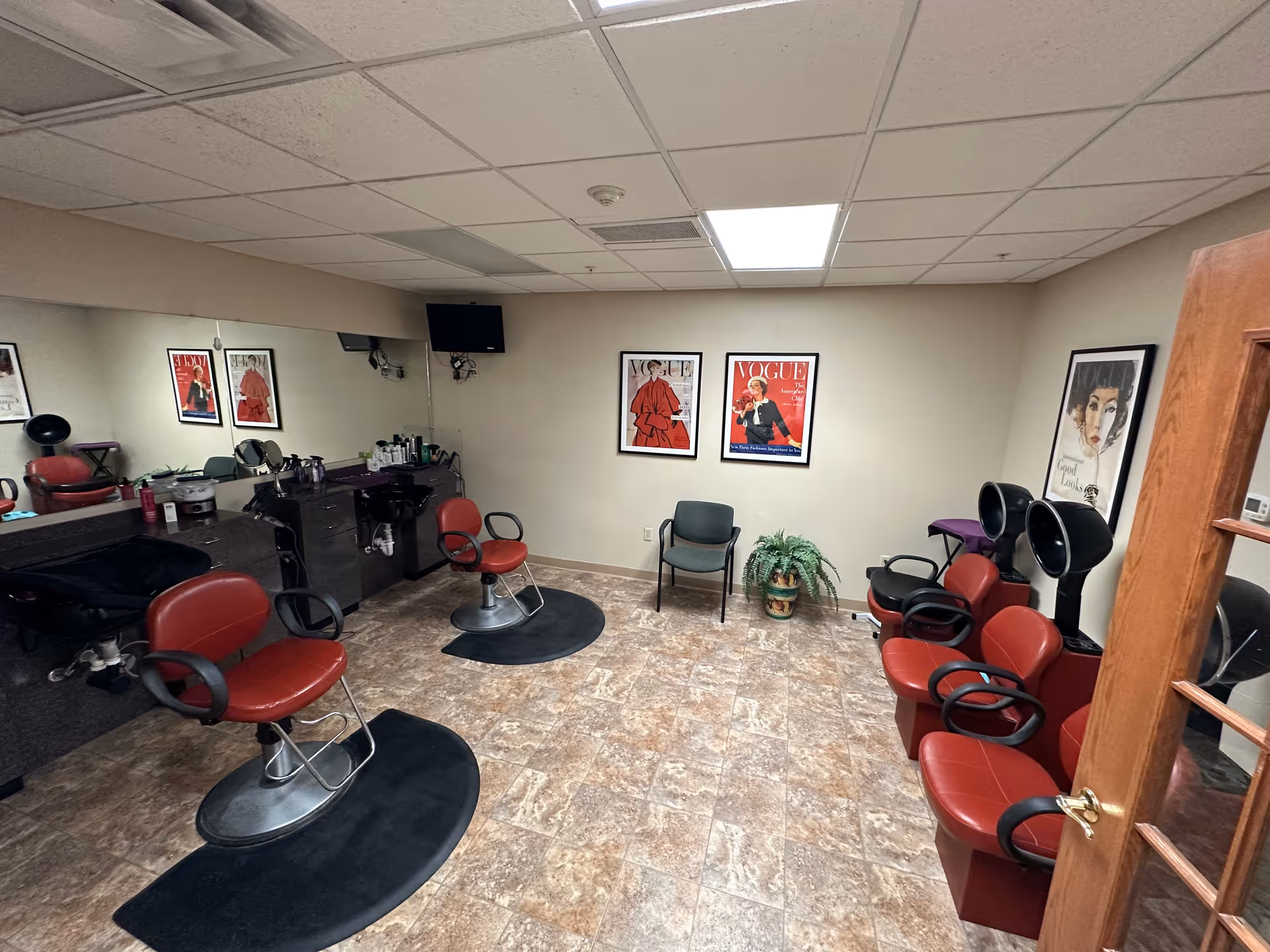 Interior salon room with red styling chairs, hooded hair dryers, mirrors and framed Vogue posters on the wall.