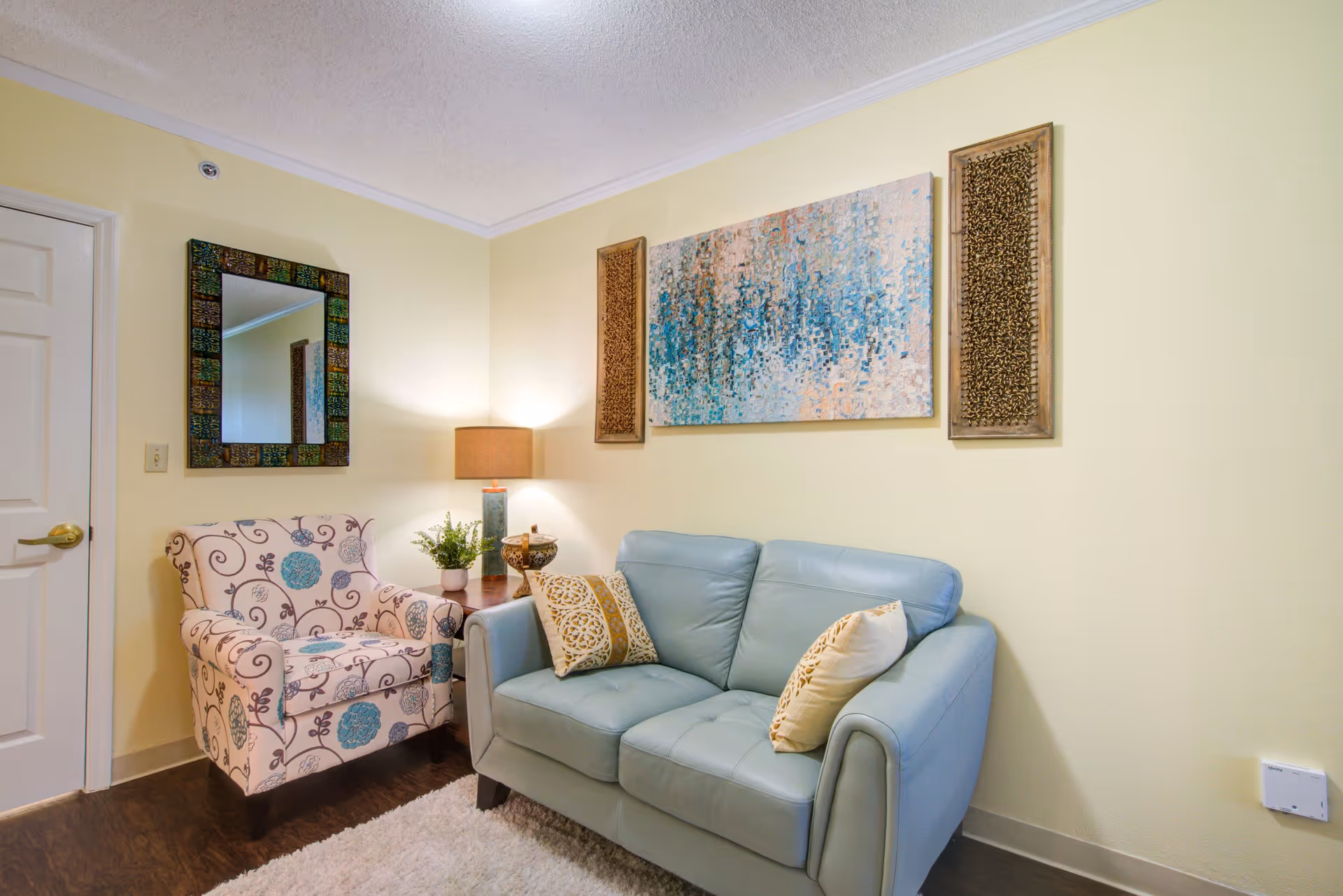 A cozy living room corner with a light blue leather loveseat adorned with two decorative pillows, a patterned armchair, a wooden side table with a lamp and a small plant, a colorful abstract painting and two vertical wooden art pieces on a pale yellow wall, and a decorative mirror hanging near a closed white door.