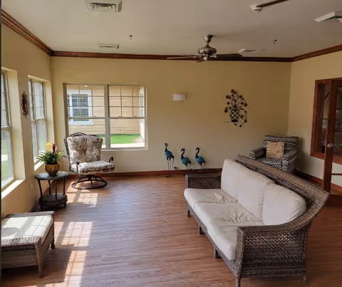 A bright and cozy living room area with wooden flooring, a ceiling fan, and large windows letting in natural light. The room is furnished with a wicker sofa with white cushions, a patterned armchair, a small round side table with a potted plant, and decorative metal peacock sculptures near the wall. There is also a wall decoration featuring a vase with flowers.