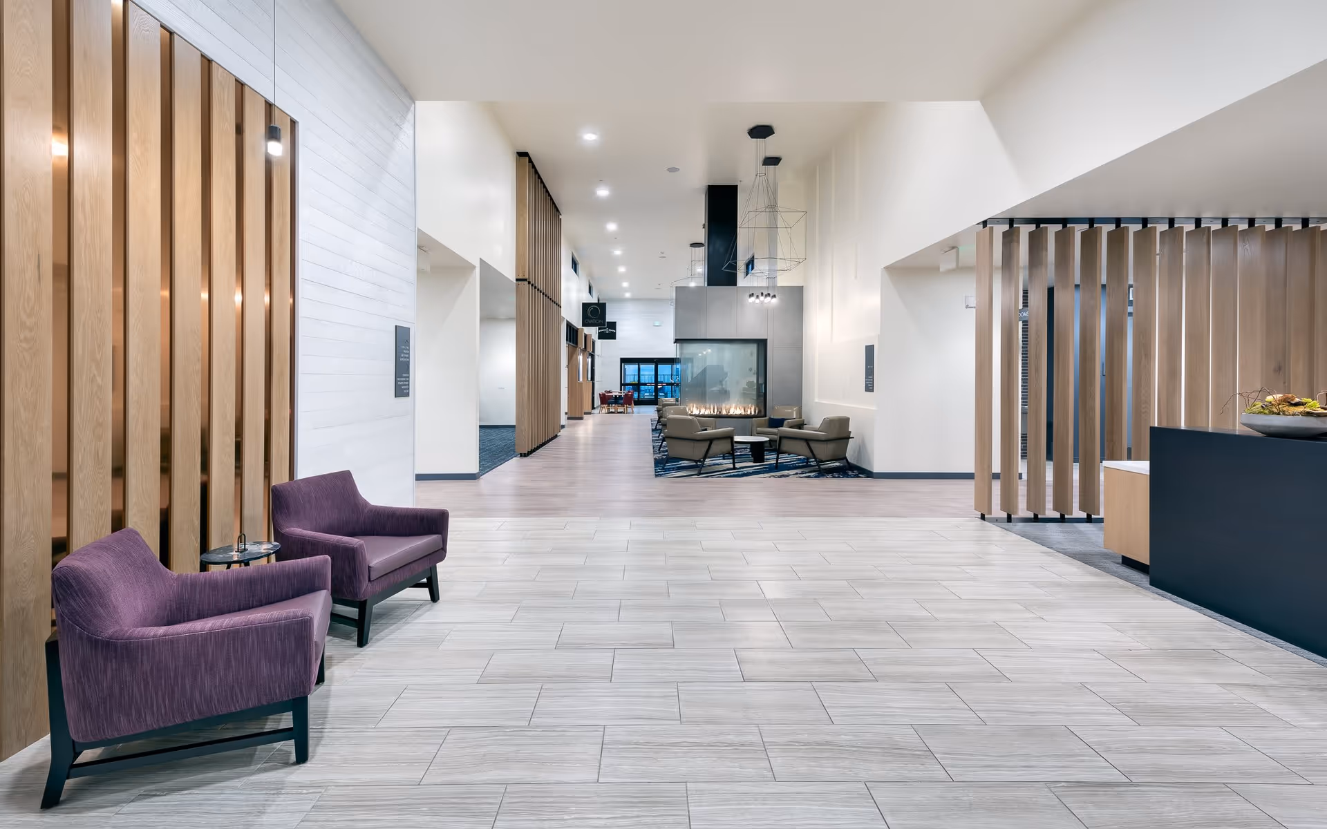 A spacious and modern interior common area with light-colored tiled flooring and white walls. Two purple armchairs are positioned on the left side near wooden vertical slats. In the background, there is a seating area with a contemporary fireplace surrounded by chairs and a rug. The space is well-lit with recessed ceiling lights and has a clean, minimalist design.