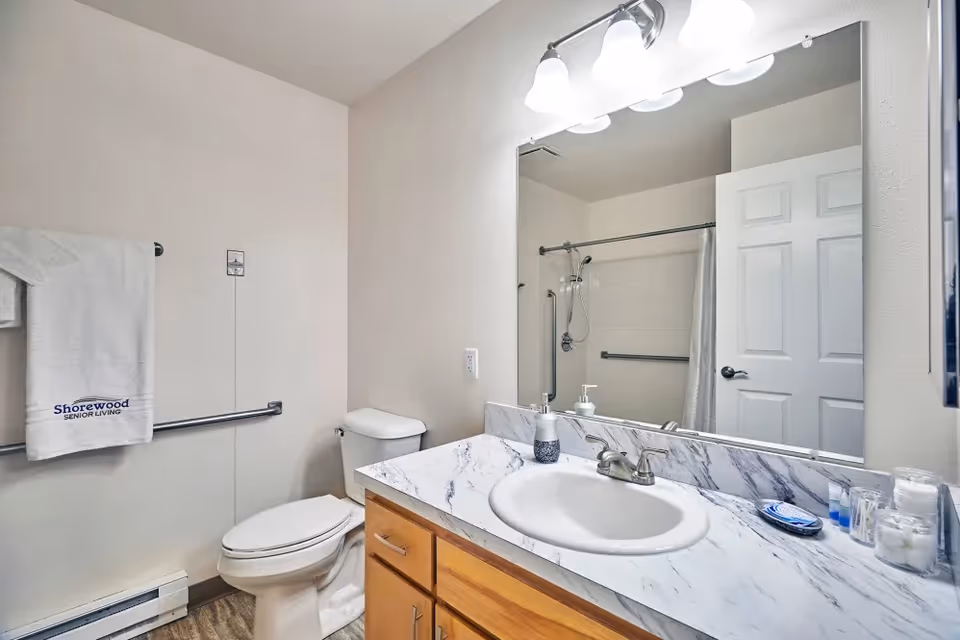 Clean bathroom with a vanity and sink, large mirror, toilet, shower, and a towel embroidered 'Shorewood SENIOR LIVING'.