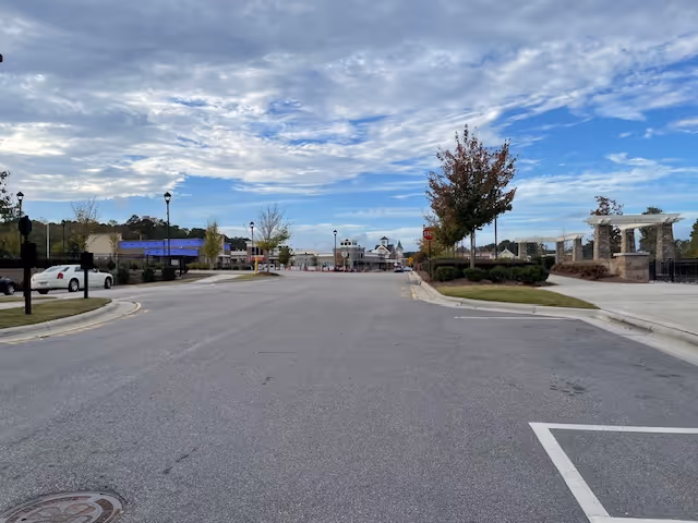 Wide empty paved street leading to a landscaped community entrance with lamp posts, trees and a pavilion under a partly cloudy sky.