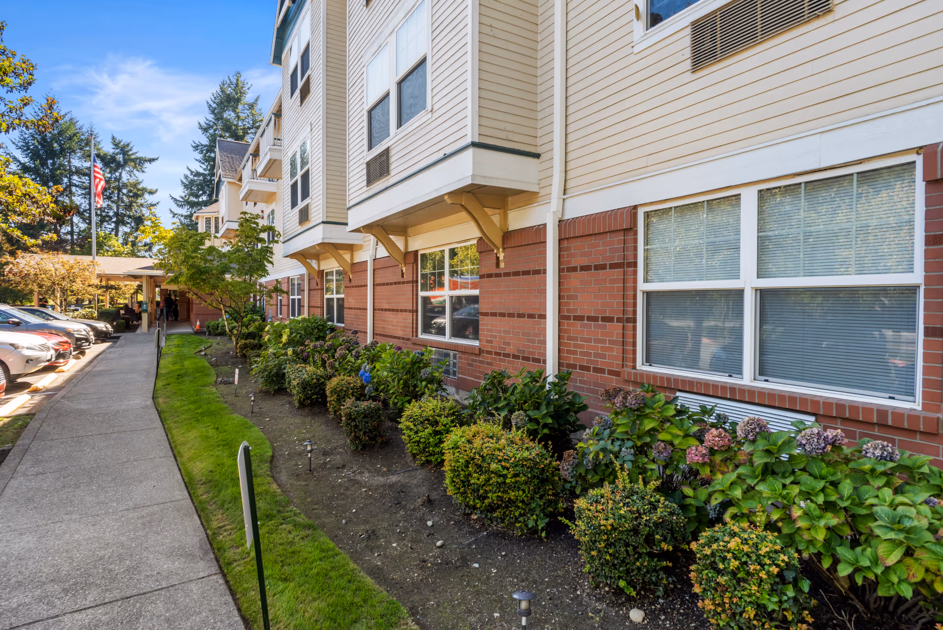 Exterior view of Bridgeport Place Assisted Living showing a sidewalk alongside a building with beige siding and red brick lower walls. There are neatly trimmed bushes and plants along the building, parked cars on the left, and an American flag in the background under a clear blue sky.