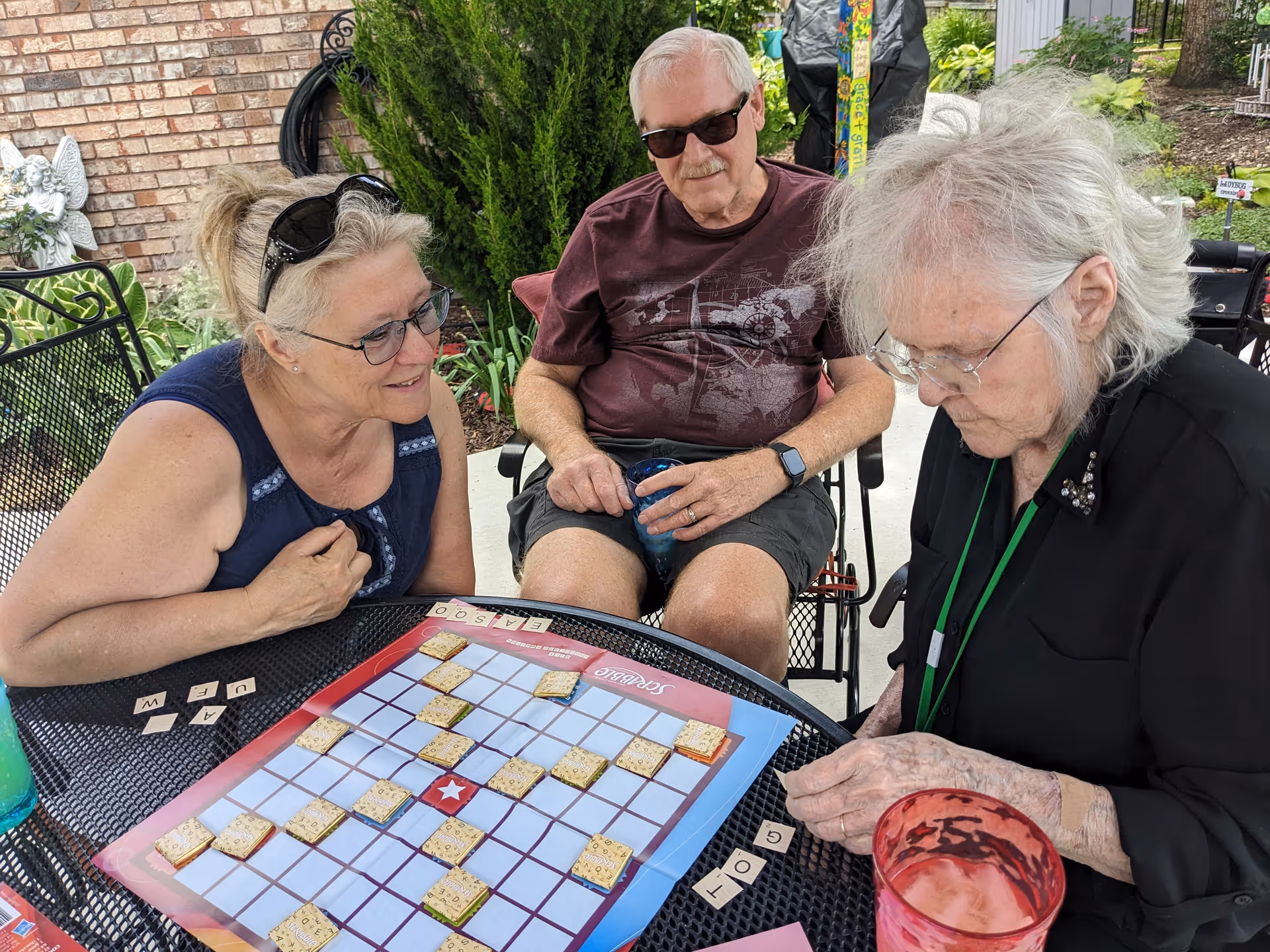Three elderly people sitting outdoors around a metal table playing a board game with letter tiles. One woman on the left is smiling and leaning forward, a man in the middle wearing sunglasses is holding a blue cup, and another woman on the right is concentrating on the game. There are plants and a brick wall in the background.