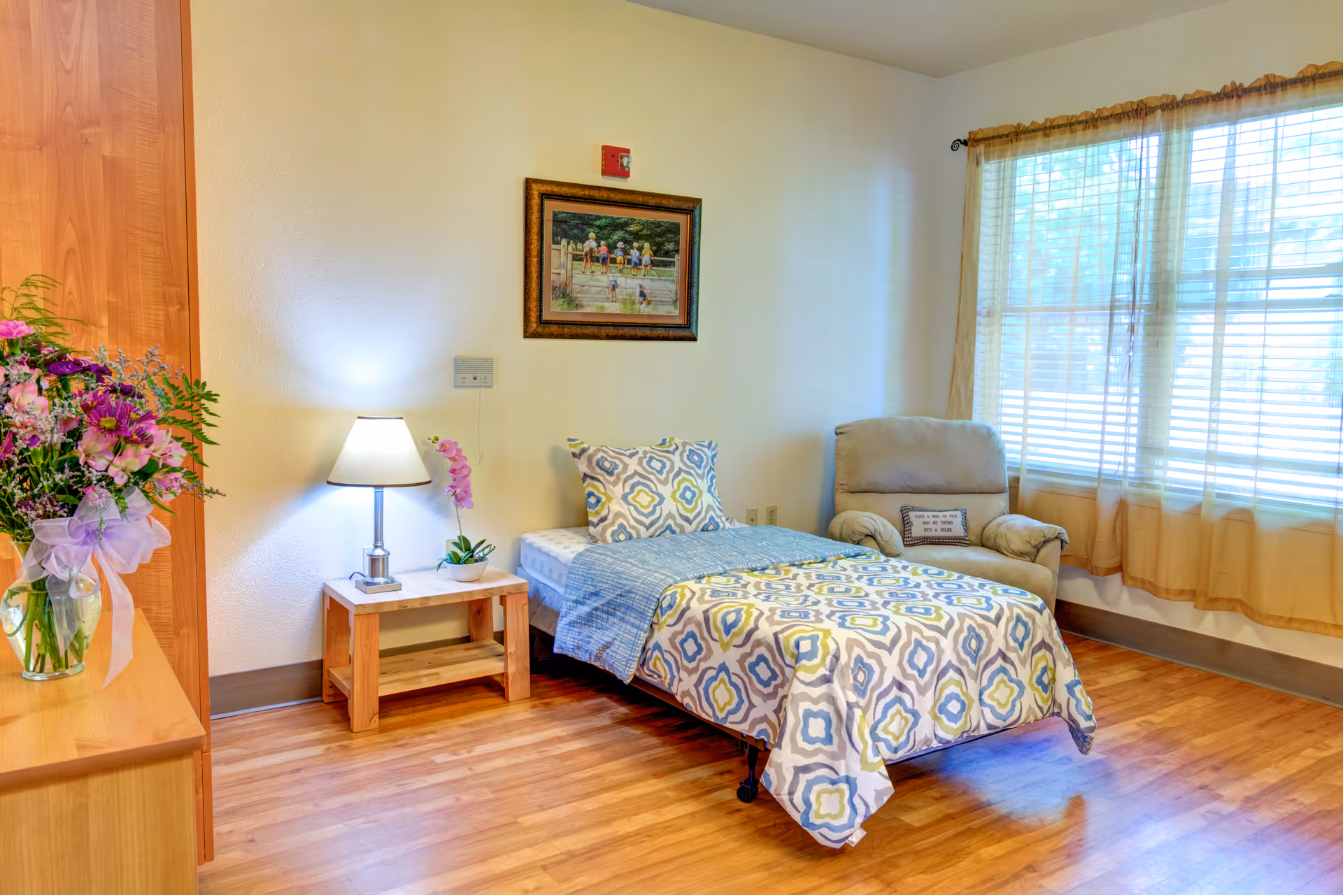 A cozy bedroom in Monterey Court Memory Care featuring a single bed with patterned bedding, a wooden nightstand with a lamp and a small plant, a comfortable armchair with a decorative pillow, a large window with sheer curtains allowing natural light to fill the room, and a framed painting on the wall.