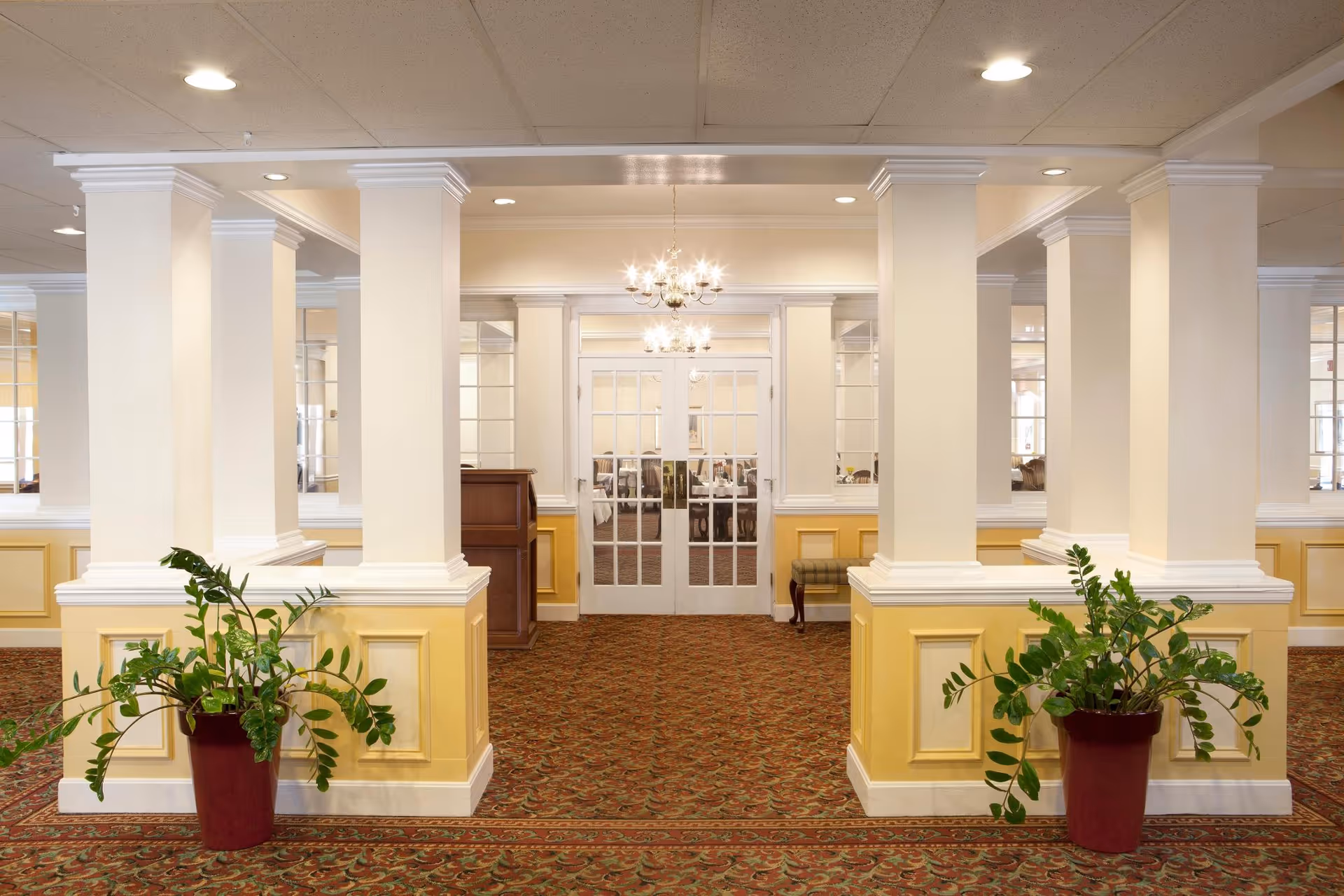 A bright interior foyer with columns, patterned carpet, potted plants, chandeliers, and double glass doors.