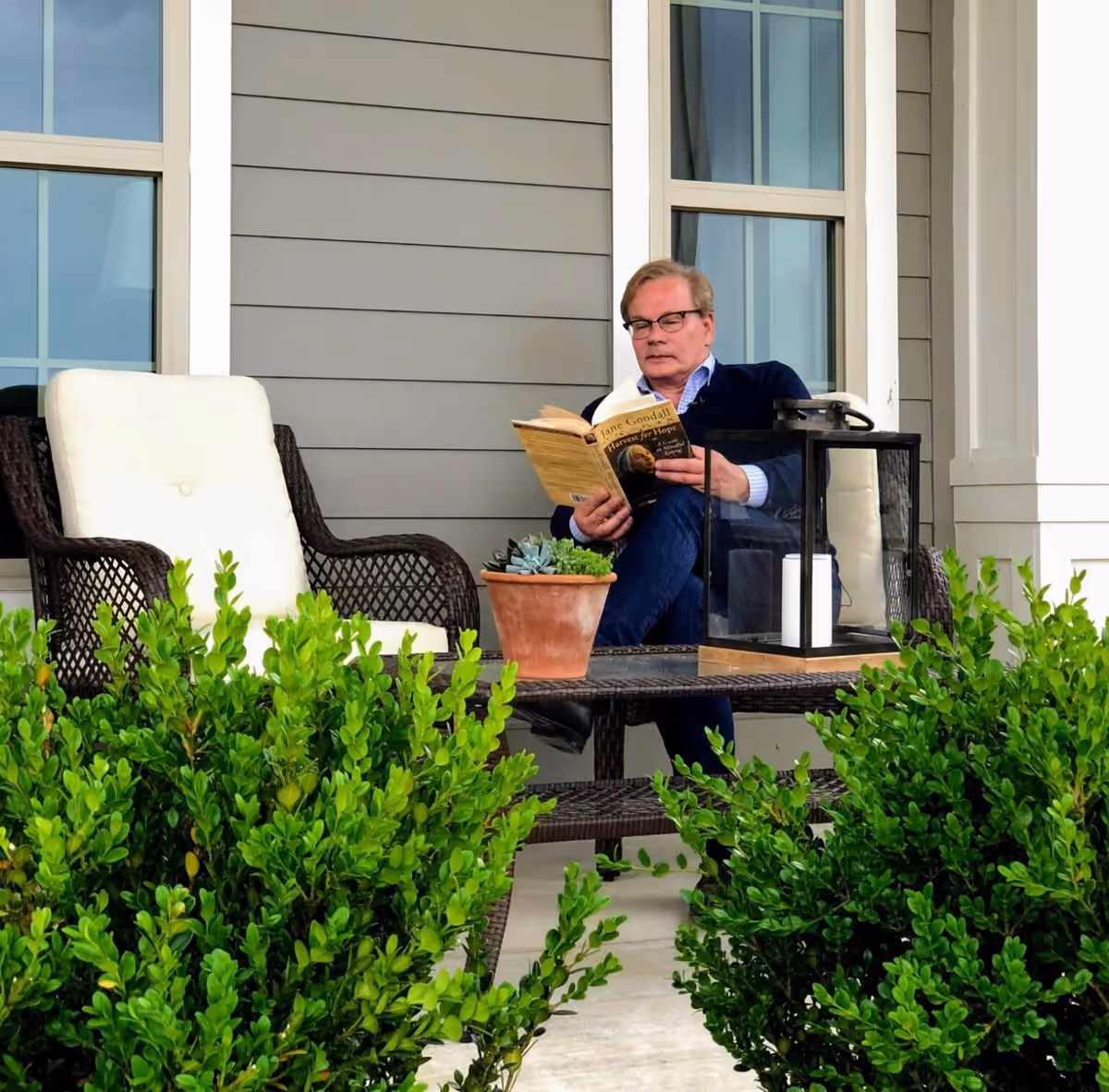 A man wearing glasses and a navy sweater is sitting on a cushioned wicker chair on a porch, reading a book titled 'Jane Goodall: Harvest for Hope'. There is a matching empty chair beside him, a wicker table with a potted plant and a lantern with candles, and green bushes in the foreground. The porch has gray siding and large windows.