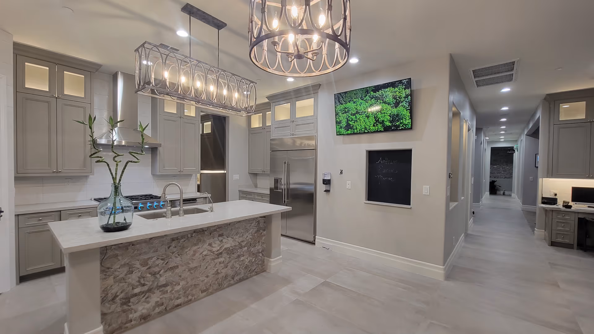 Modern kitchen area with a large island featuring a stone facade and a glass vase with bamboo stalks. The kitchen has gray cabinets, a stainless steel refrigerator, and a stove with a range hood. Two decorative pendant lights hang above the island. A flat-screen TV is mounted on the wall above a small chalkboard with the words 'Anthem Senior Manor' written on it. A hallway extends to the right with additional rooms and a desk area visible.