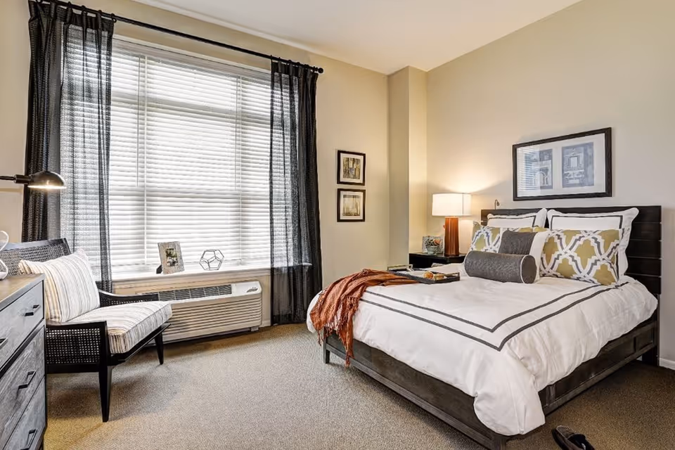 A well-lit bedroom with a large window covered by sheer black curtains and white blinds. The room features a neatly made bed with white and patterned pillows and a brown throw blanket. There is a bedside table with a lamp and decorative items, a cushioned chair with striped upholstery, and a dresser with a lamp. The walls are painted beige and decorated with framed artwork.