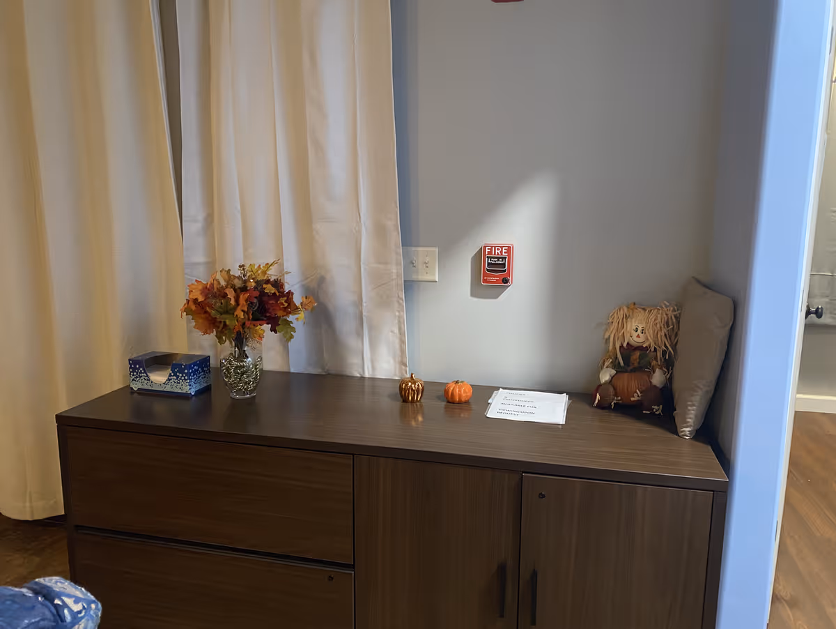 A wooden cabinet against a light gray wall with a fire alarm mounted above it. On the cabinet are a vase with autumn leaves, two small decorative pumpkins, a tissue box, a piece of paper, and a small doll with yarn hair sitting next to a pillow. Beige curtains hang to the left side of the cabinet.