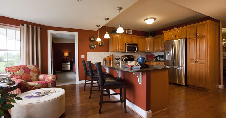 Interior view of a kitchen and living area in a senior living facility. The kitchen features wooden cabinets, a stainless steel refrigerator, microwave, and a countertop with three black bar stools. The living area has a patterned armchair and a round ottoman with magazines on it. A doorway leads to a bedroom with a nightstand and lamp visible. The walls are painted a warm reddish-brown color, and the floor is wooden.