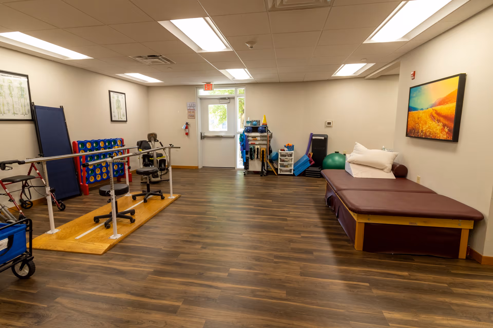 A rehabilitation or physical therapy room with wooden flooring, parallel bars for walking exercises, chairs on wheels, a large Connect Four game, exercise equipment, a padded therapy table with pillows, and a colorful landscape painting on the wall.