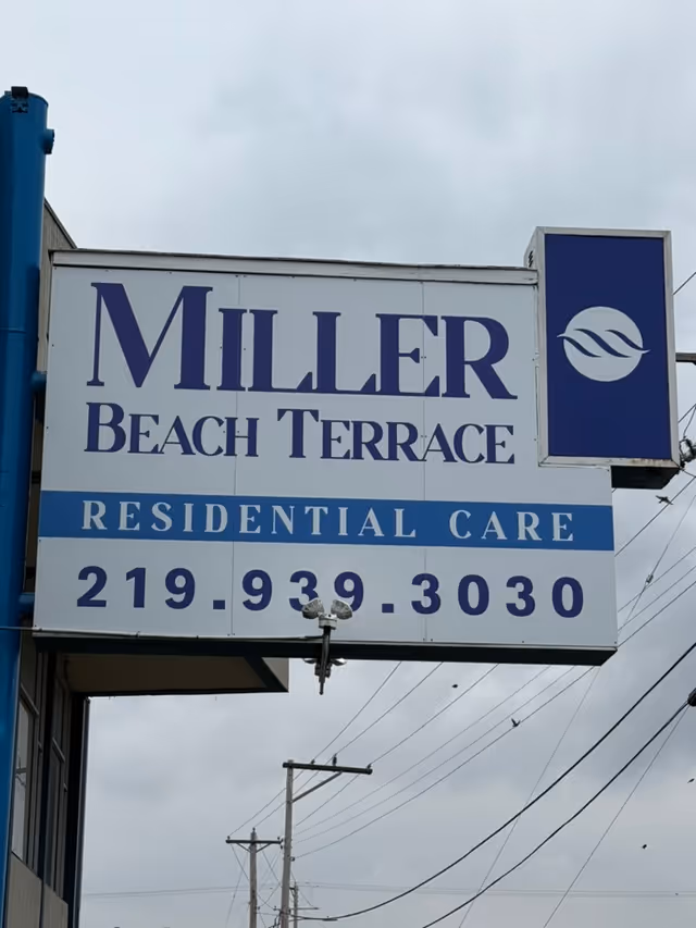 Outdoor sign reading "Miller Beach Terrace Residential Care" with a phone number mounted on a building against a cloudy sky.