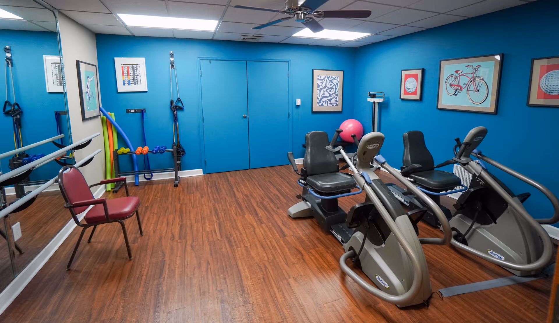 Small fitness room with two recumbent exercise bikes, a mirror, resistance bands and dumbbells against blue walls and wood flooring.