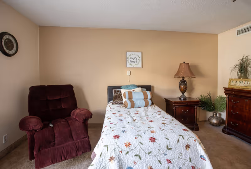 A cozy bedroom with a single bed covered in a floral quilt, several decorative pillows, a dark red recliner chair to the left, a wooden nightstand with a decorative lamp, and a wooden dresser with a plant and a 'FAMILY' sign on top. A clock hangs on the left wall and a framed sign that reads 'simply blessed' is centered above the bed.