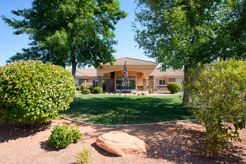 Front exterior view of a single-story assisted living facility building with a covered entrance, an American flag on a flagpole in the center, surrounded by green grass, trees, and shrubs under a clear blue sky.