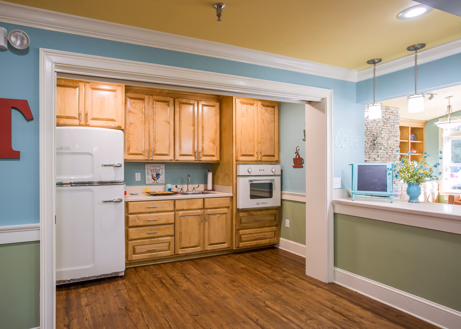 A cozy kitchen area with wooden cabinets, a white vintage-style refrigerator, a built-in oven, and a sink. The walls are painted blue and green with white trim, and the ceiling is yellow. There is a counter with a vase of flowers and a small retro-style television on it. Pendant lights hang from the ceiling, and a stone accent wall is visible in the adjoining room.