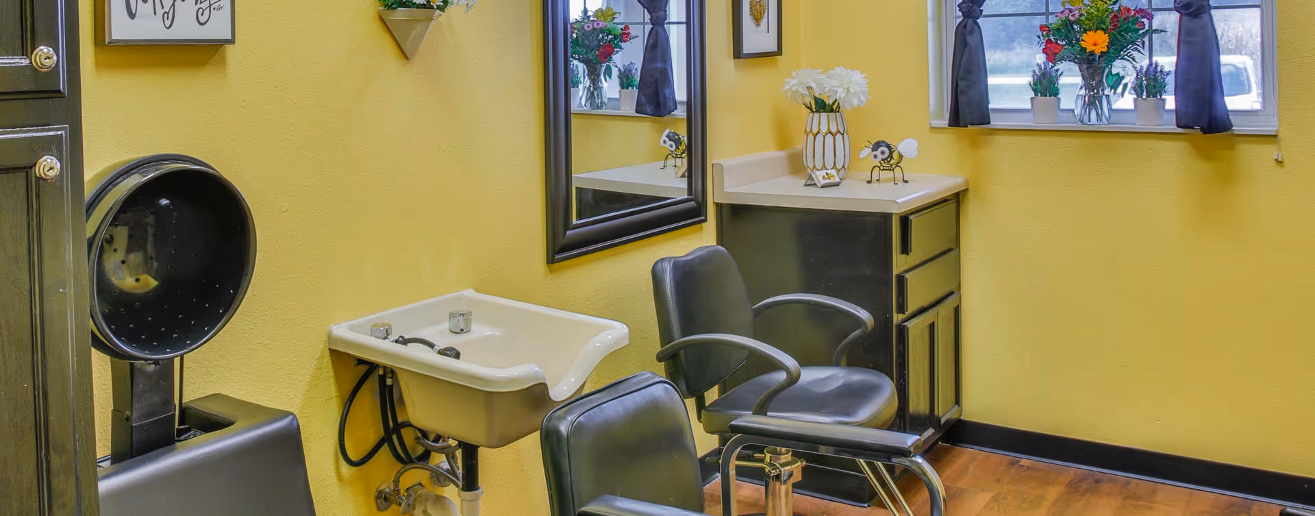 Interior of a small salon area with two black salon chairs, a white sink for washing hair, a large mirror on the yellow wall, a window with dark curtains, and decorative flowers on the countertop and window sill.