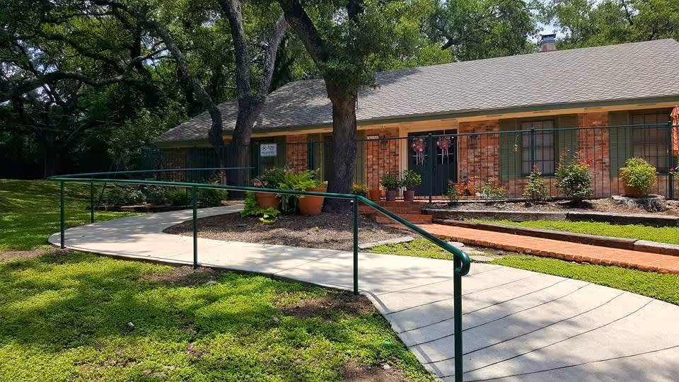 A single-story brick building with green shutters and a green door, surrounded by trees and plants. A concrete walkway with a green handrail curves towards the entrance, and there are flower pots and small bushes near the door.
