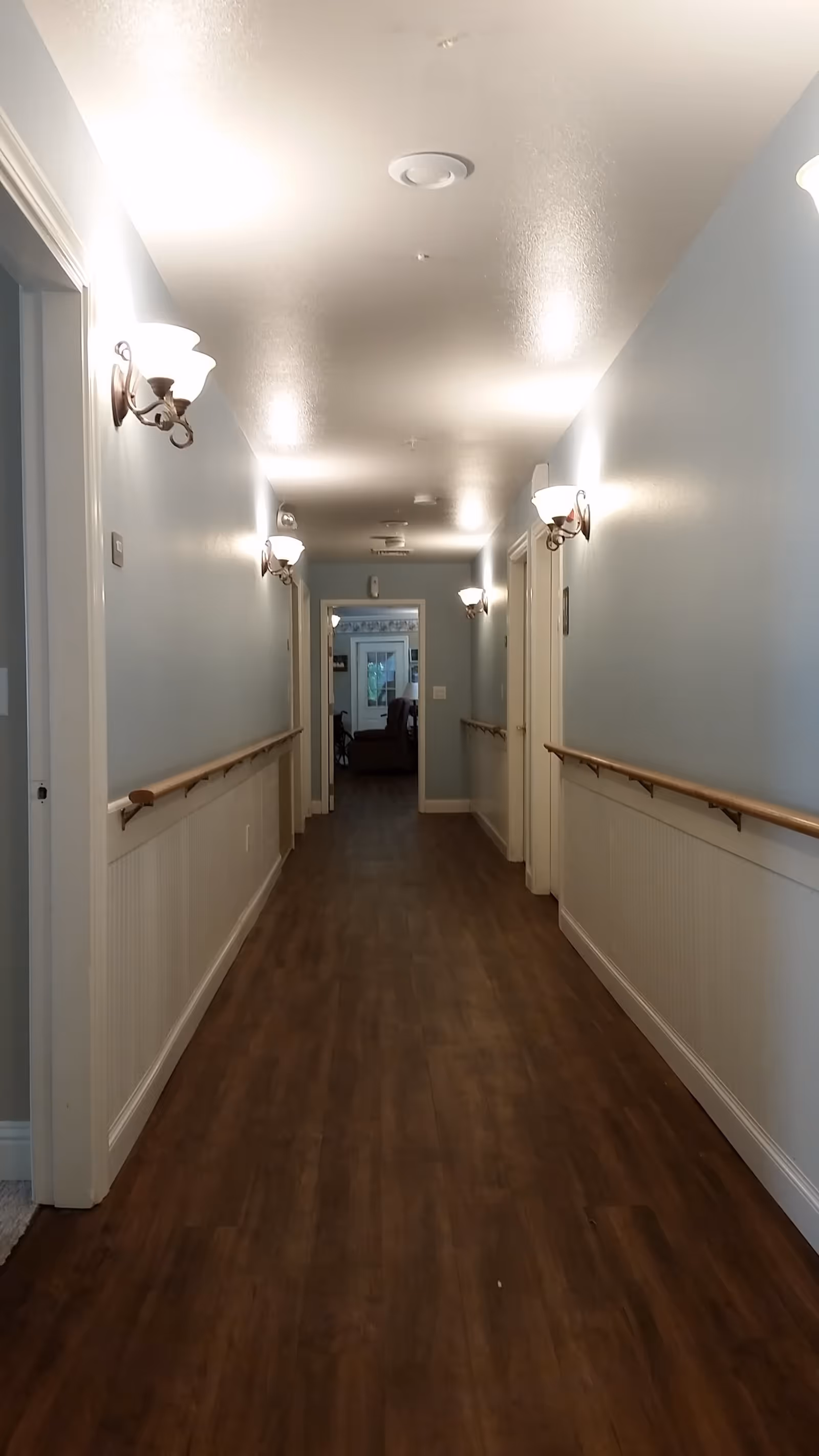 A long, well-lit hallway with wooden flooring and light blue walls. The hallway has handrails on both sides and several wall-mounted light fixtures. Doors line both sides of the hallway, and at the end, there is an open doorway leading to another room with a window and furniture visible.