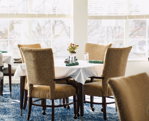 A dining area with a round table covered with a white tablecloth, surrounded by four upholstered chairs with patterned fabric. The table is set with green napkins, glasses, and a small vase of flowers. Large windows in the background let in natural light.