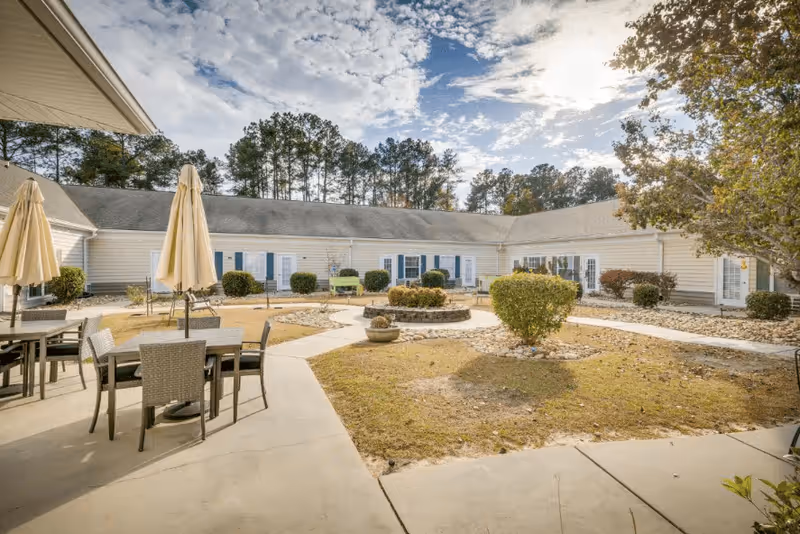 Outdoor courtyard area of a senior living facility with patio tables and chairs under closed umbrellas, surrounded by a single-story building with white siding and blue shutters. There are bushes, a small circular stone planter, and trees in the background under a partly cloudy sky.