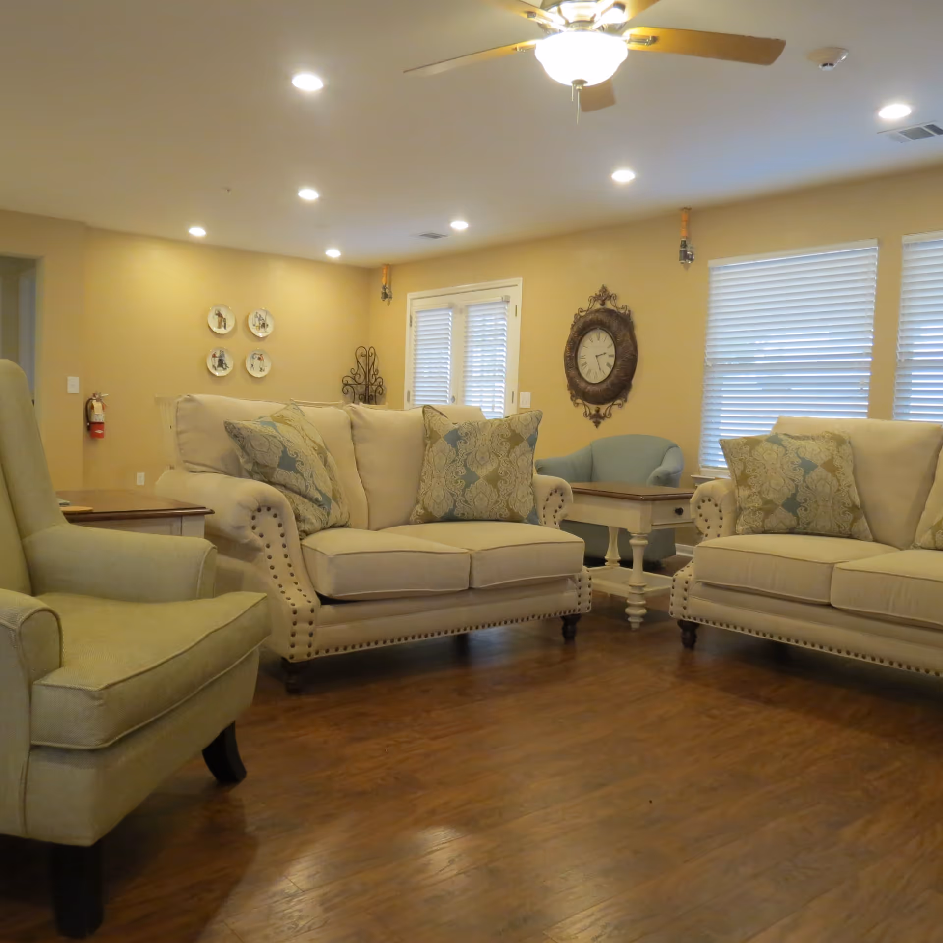 A cozy living room with beige walls and wooden flooring featuring two beige sofas with patterned cushions, a beige armchair, a small wooden side table, a blue armchair, a wall clock, and windows with blinds. The ceiling has recessed lighting and a ceiling fan with wooden blades.