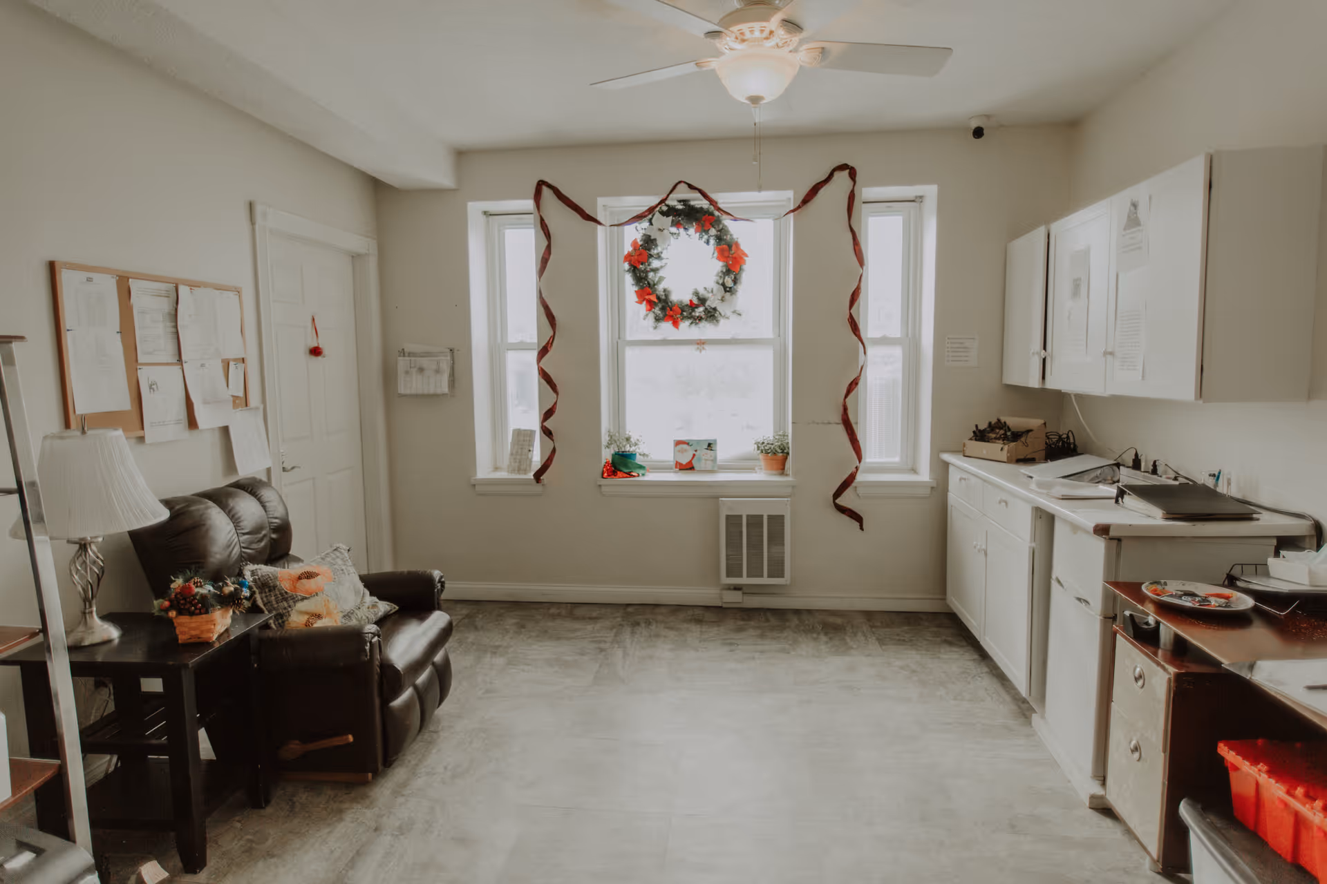 A cozy room with a brown leather loveseat, a side table with a lamp and a small basket of flowers, a bulletin board with papers on the wall, and a window decorated with a Christmas wreath and red ribbons. On the right side, there are white cabinets, a countertop with various items, and a small heater below the window.