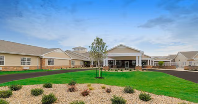Front exterior view of West River Health Campus showing a single-story building with beige siding and stone accents, a covered entrance with white columns, a circular driveway, and landscaped greenery including grass, small bushes, and a young tree under a partly cloudy sky.