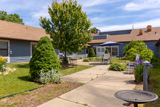 Outdoor courtyard area at a senior living facility with a concrete walkway, green grass, various shrubs and trees, white chairs, and a table with a blue umbrella under a partly cloudy sky.