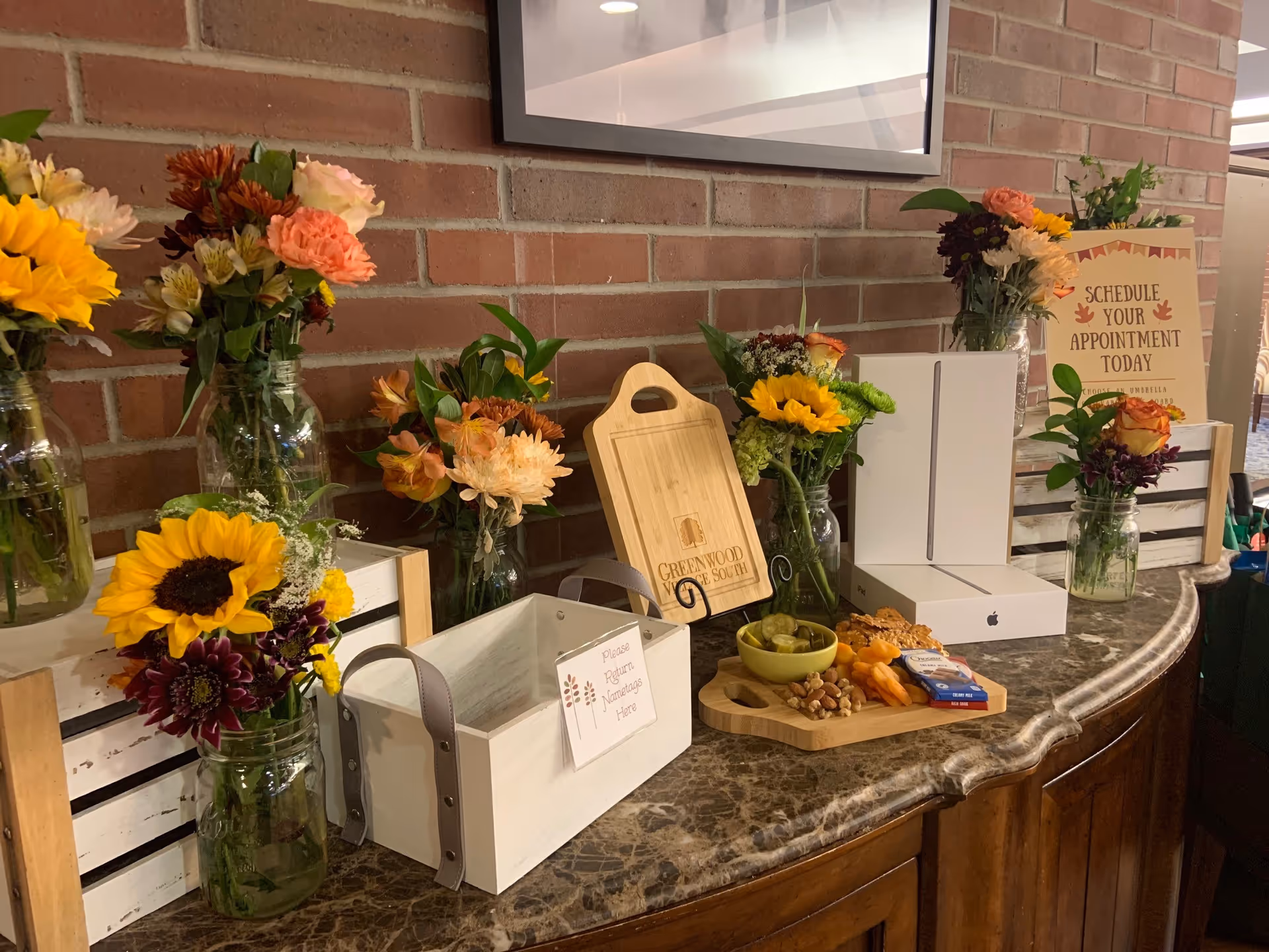 A marble countertop against a brick wall decorated with multiple glass jars filled with colorful flowers including sunflowers, roses, and daisies. On the countertop, there is a wooden cutting board engraved with 'Greenwood Village South,' a white wooden box with a sign that says 'Please Return Nametags Here,' a small bowl with pickles, nuts, and dried apricots on a wooden tray, and a boxed Apple iPad. A sign in the background reads 'Schedule Your Appointment Today.'