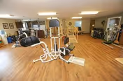 A spacious fitness room with wooden flooring featuring various exercise equipment including a multi-station gym machine, stationary bikes, and other workout apparatus. The room has beige walls, several windows, and a staircase leading to an upper level.