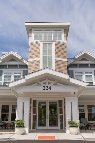 Front exterior view of a senior living facility named The Residence at Vinnin Square, featuring a large entrance with double glass doors, white columns, beige siding, and the number 224 above the entrance. There are potted plants on either side of the entrance and a clear sky in the background.