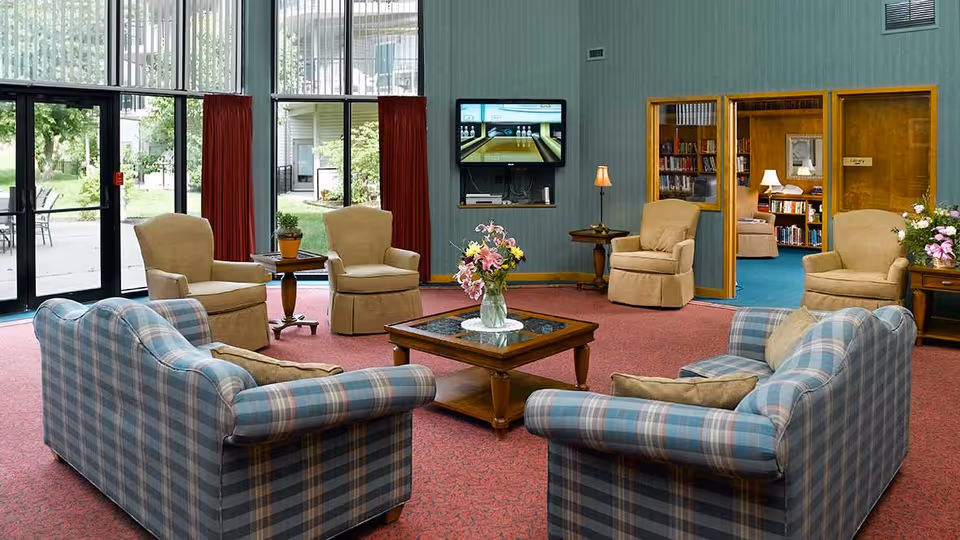 A cozy living room area with two blue plaid sofas and four beige armchairs arranged around a wooden coffee table with a vase of flowers. Large windows with red curtains let in natural light and show an outdoor patio area. A TV is mounted on the wall, and a doorway leads to a library room filled with bookshelves and more seating.