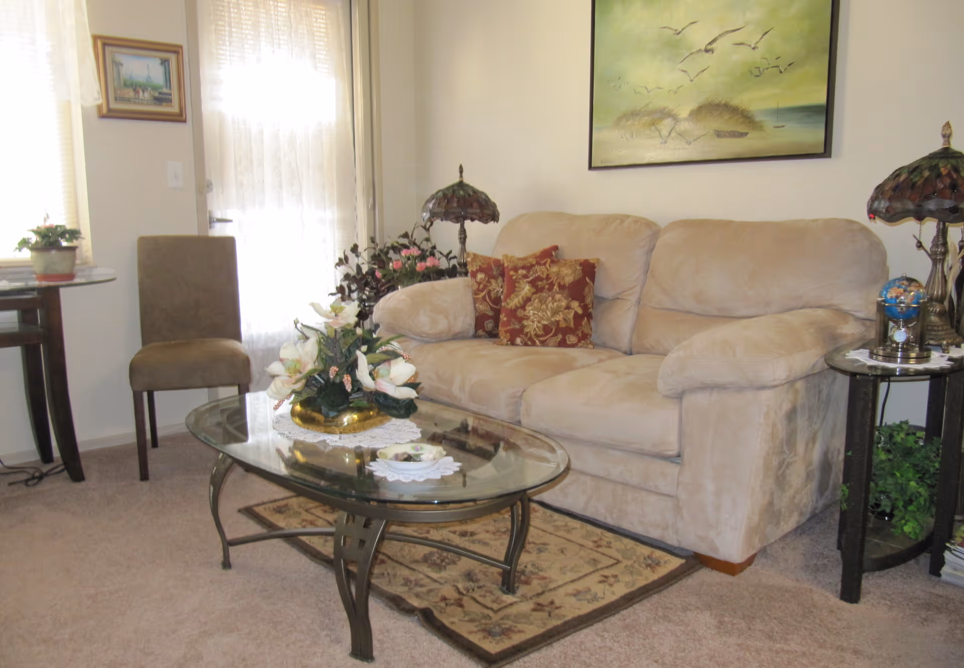 Cozy living room with a beige loveseat, glass coffee table holding a floral centerpiece, side tables, and wall art.