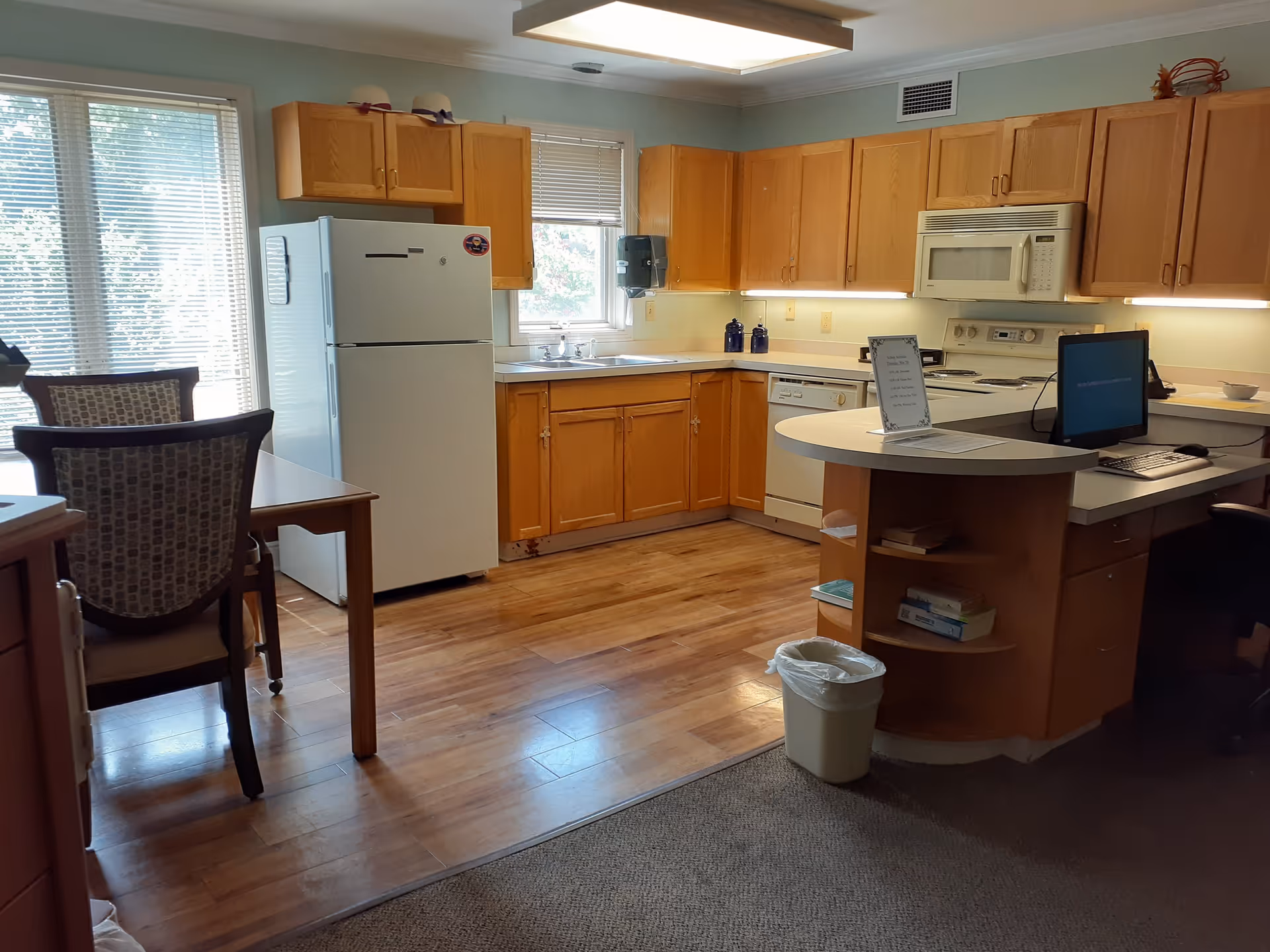 Interior view of a kitchen area with wooden cabinets, a white refrigerator, a microwave, a stove, a dishwasher, and a sink under a window. There is a curved countertop with a computer monitor and keyboard on it. To the left, there is a dining table with chairs near large windows with blinds.