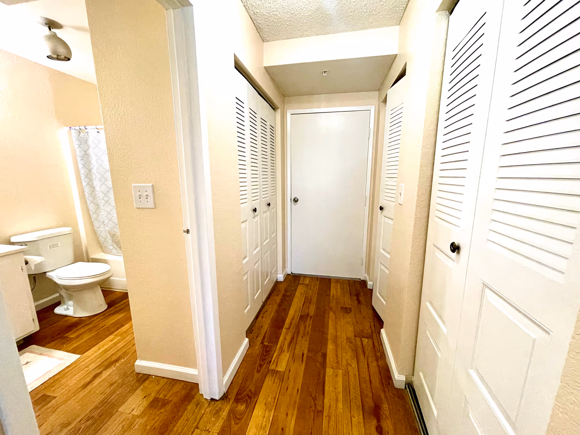 A hallway with wooden flooring and beige walls leading to a white door at the end. On the left side, there is an open doorway showing a bathroom with a toilet, sink, and a shower curtain. Both sides of the hallway have white louvered closet doors.