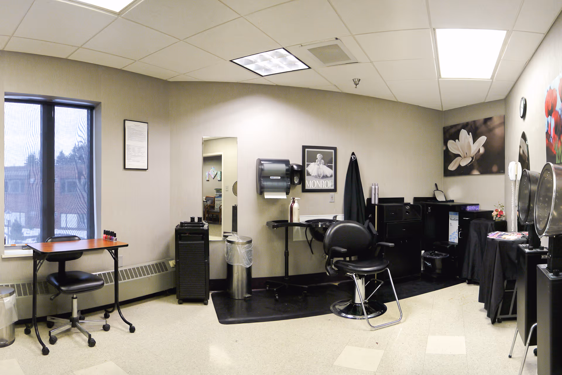 Interior view of a salon room in a senior living facility with a styling chair in front of a hair washing station, a mirror, a small table with a chair near a window, and various salon equipment and decorations on the walls.