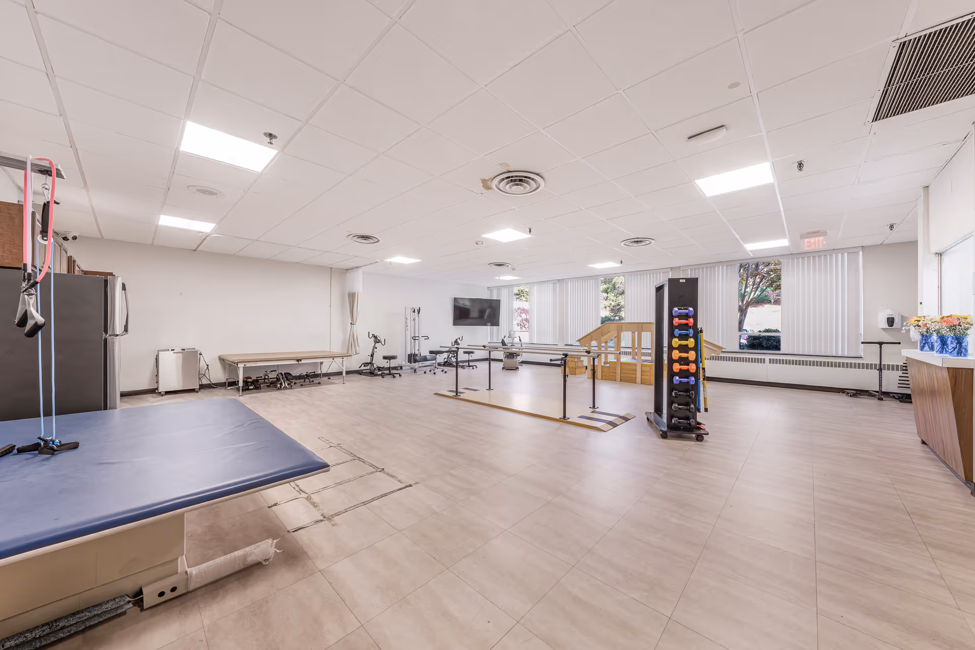 A spacious physical therapy or rehabilitation room with exercise equipment including parallel bars, a padded therapy table, a rack of colorful dumbbells, and a small wooden ramp with handrails. The room has large windows with vertical blinds, light-colored tiled flooring, and a white ceiling with recessed lighting.