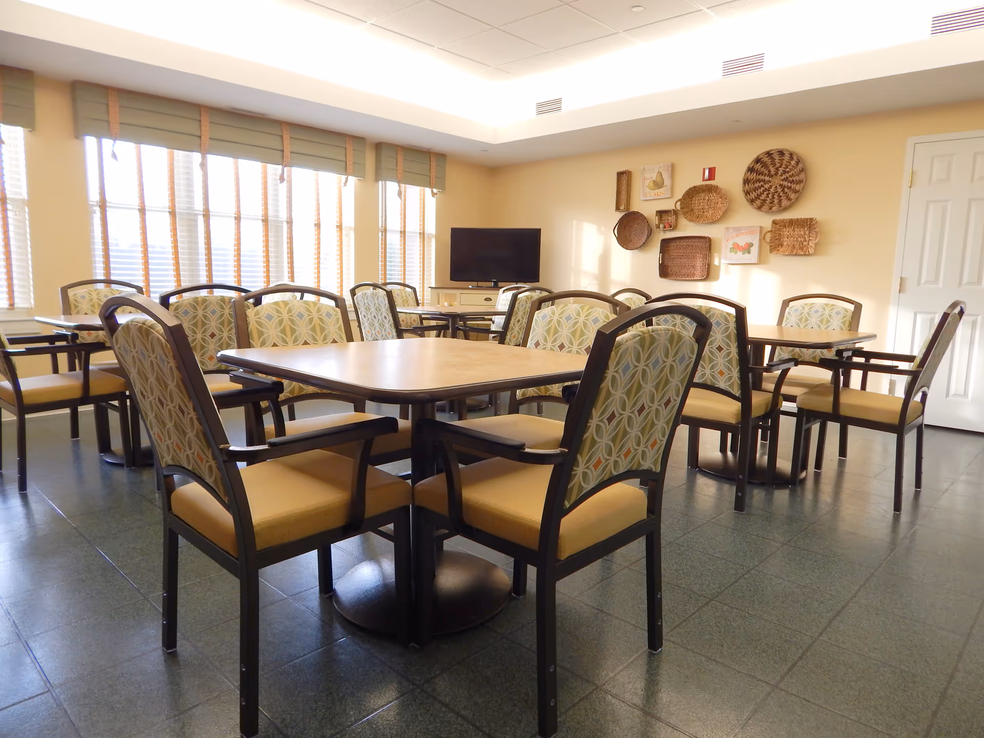 A bright dining room with several tables and cushioned chairs arranged neatly. Large windows with blinds allow natural light to fill the room. A flat-screen TV is mounted on a cabinet against the wall, which is decorated with various woven baskets and framed pictures.