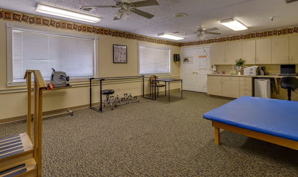 A therapy or rehabilitation room with exercise equipment including a stationary pedal exerciser, parallel bars for walking support, a padded therapy table, and a small set of stairs with handrails. The room has beige walls, carpeted floor, two windows with blinds, ceiling fans, fluorescent lighting, and cabinets with a countertop holding various items including a microwave and a small refrigerator.