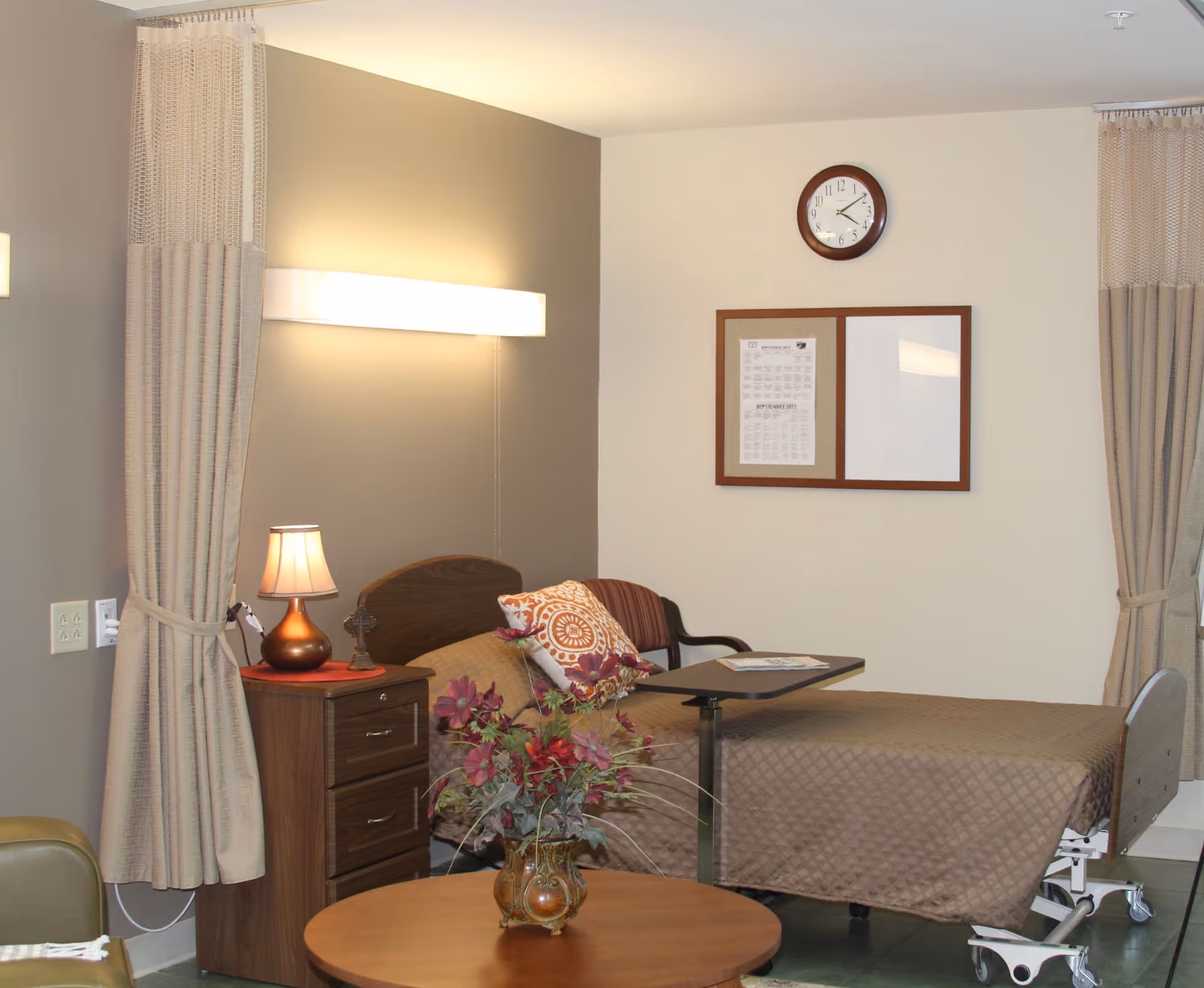 A nursing facility room with a single hospital bed covered in a brown quilt, a bedside table with a lamp and a small cross, a chair with a decorative pillow, a round wooden table with a vase of flowers, beige curtains, a wall clock, and a bulletin board on the wall.