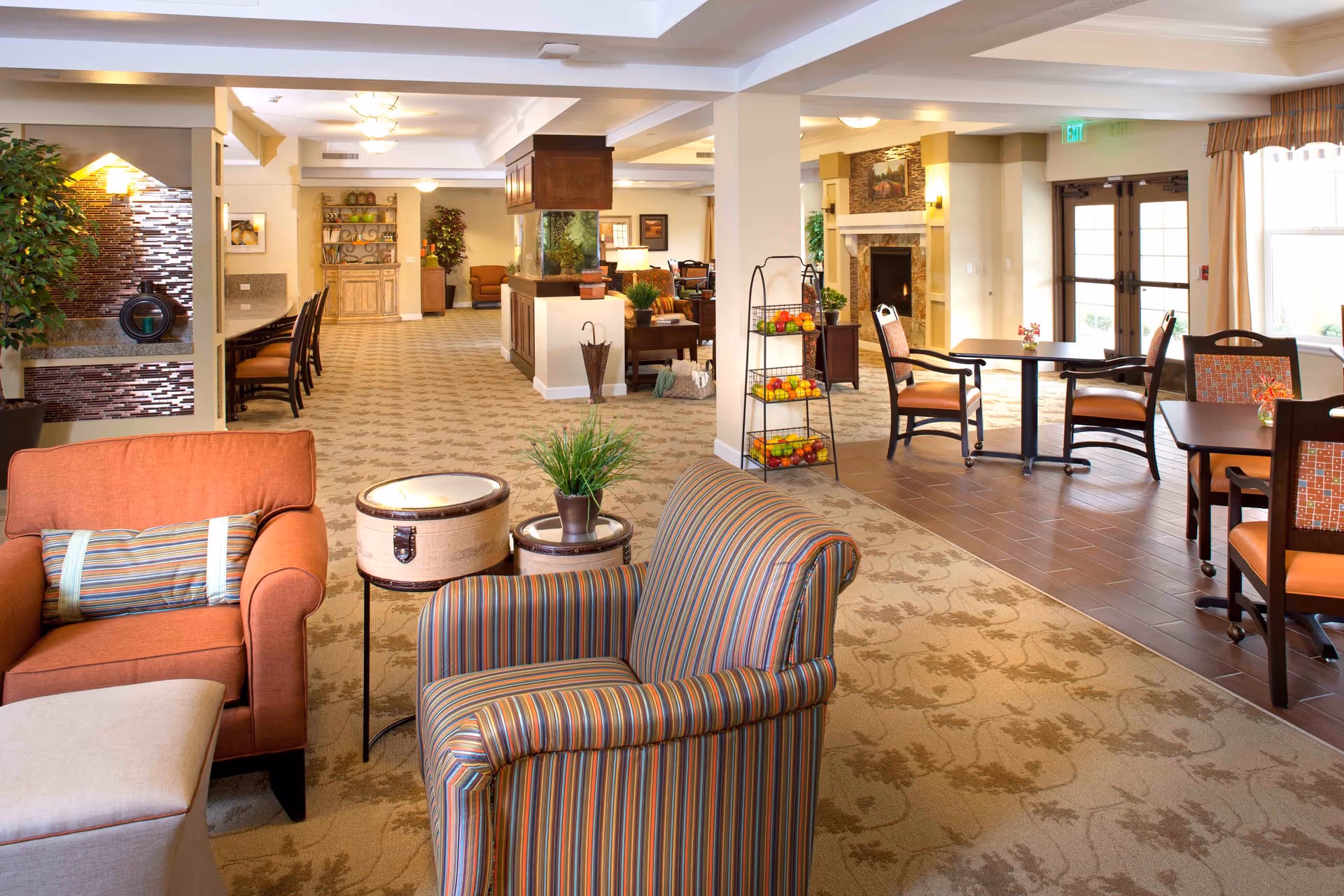 Spacious, well-lit common area with upholstered chairs, tables, a fireplace and decorative plants in an assisted living facility.