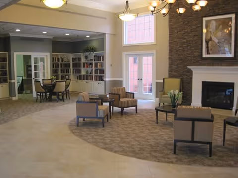 A spacious senior living common area with a mix of tiled and carpeted flooring. The room features several armchairs and small tables arranged for conversation, a stone fireplace with a framed artwork above it, and large windows letting in natural light. In the background, there is a dining area with a round table and chairs, built-in bookshelves, and a doorway leading to another room.