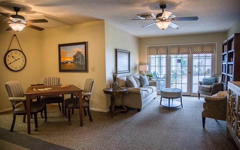 A cozy living room and dining area in a senior living facility. The dining area has a wooden table with four chairs and a chessboard on top. A large wall clock and a framed painting hang on the wall. The living room features a beige sofa, two armchairs, a round coffee table, a side table with a lamp, and a bookshelf. Large glass doors lead to an outdoor patio area.