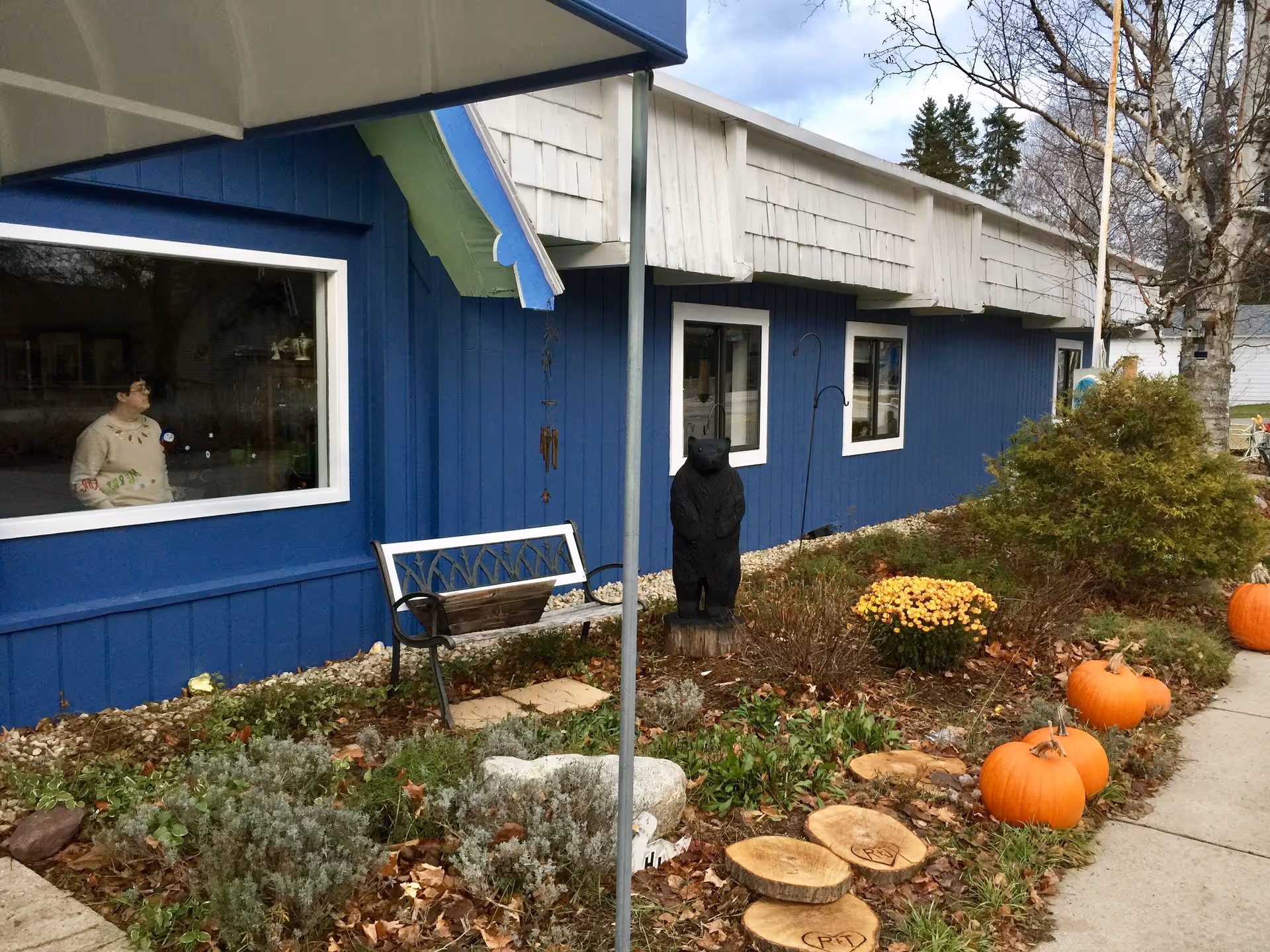 Exterior view of a blue building with white trim, featuring a garden bed with pumpkins, flowers, and shrubs. A wooden bench and a carved wooden bear statue are placed near the building. A person is visible inside the building through a large window.