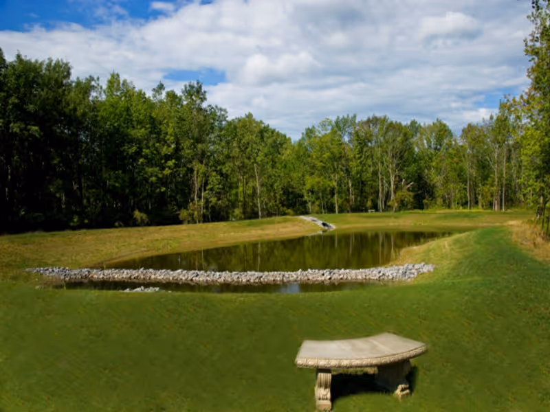 Small ornamental pond surrounded by grassy berms and trees with a stone bench in the foreground under a partly cloudy sky.